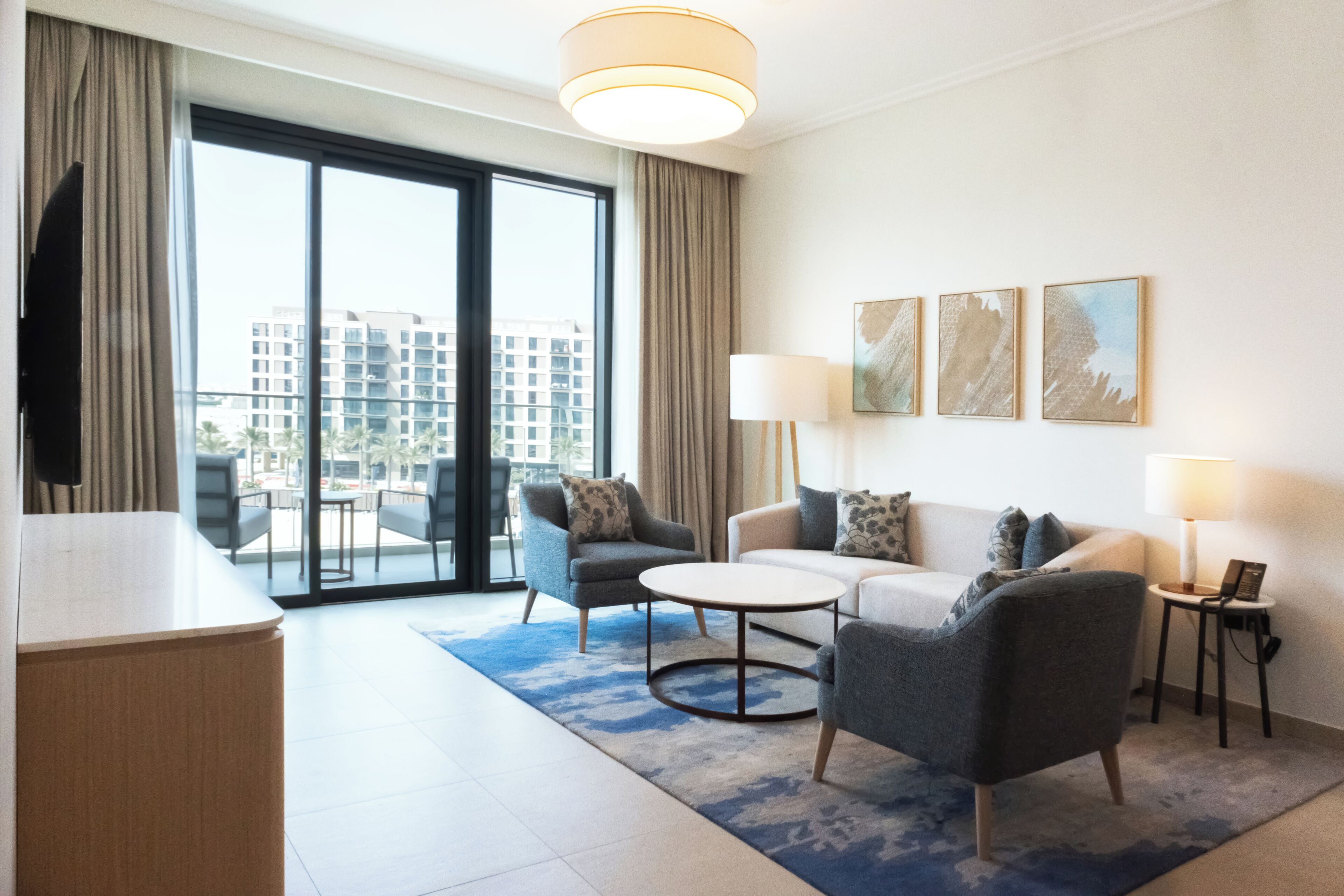 Una habitación de hotel moderna con sofá, sillones, una mesa de centro, televisión y un ventanal con vistas a la ciudad.