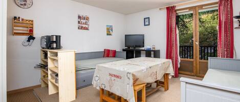 A compact living space with a kitchenette, a dining table, a TV, and a balcony with red curtains.