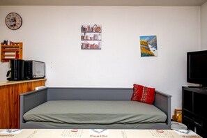 Ein Einzelbett mit grauem Kopfteil, einem roten Kopfkissen, einer Uhr, einem Regal mit Gläsern und einem Fernseher.