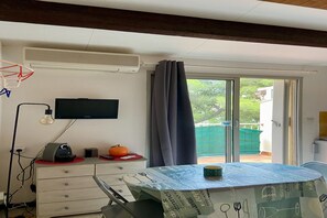 A room with a TV mounted on the wall, a table with a tablecloth, a chair, a desk with a lamp, and a window with curtains. - Bright apartment in Agde near the beach (Agde)