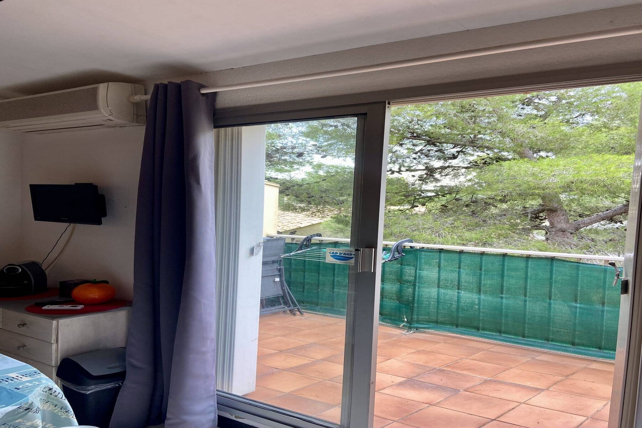 A room with a sliding glass door leading to a balcony with a green fence and trees.