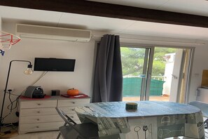 A room with a table covered with a tablecloth, a television on the wall, a ceiling fan, and a window with curtains. - Bright apartment in Agde near the beach (Agde)