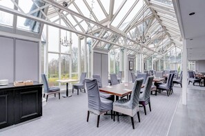 A spacious room with a high ceiling and large windows, featuring a modern dining setup with tables and chairs.