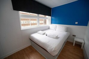 A bedroom with a bed, a window, a small bedside table, and a blue accent wall. - Small Double Room - No Window (Luton)