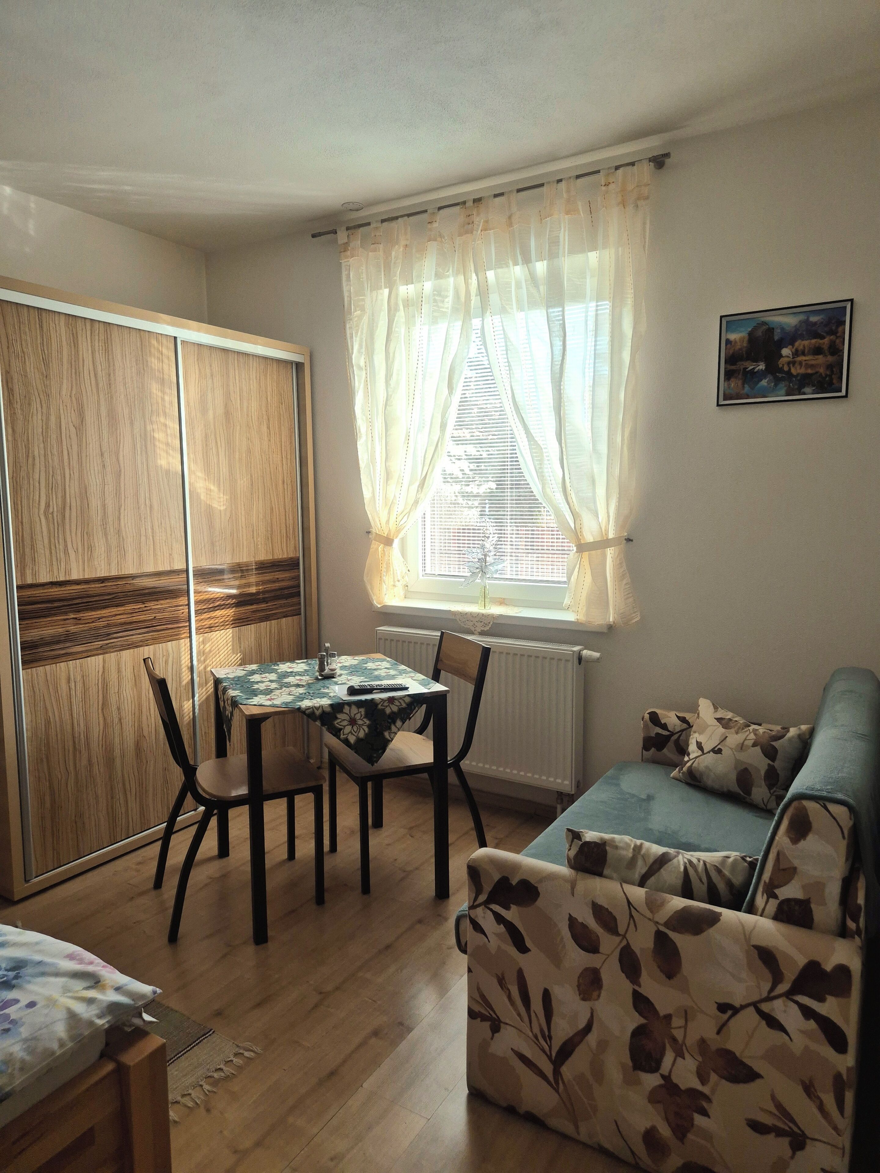 A room with a dining table, chairs, a sofa, a radiator, and a window with curtains.