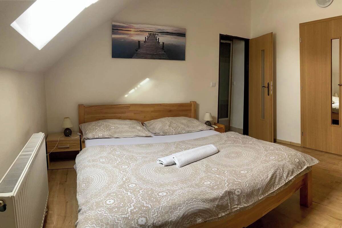 Un dormitorio con tragaluz, una cama con ropa de cama estampada, una mesita de noche con lámpara y un armario de madera.