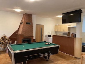 A pool table in a room with a fireplace, a kitchen area, and a TV mounted on the wall.