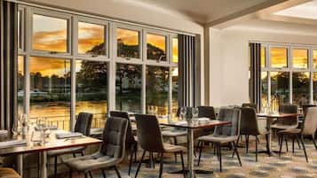 A dining area with tables and chairs, large windows with a view of a sunset over a body of water.