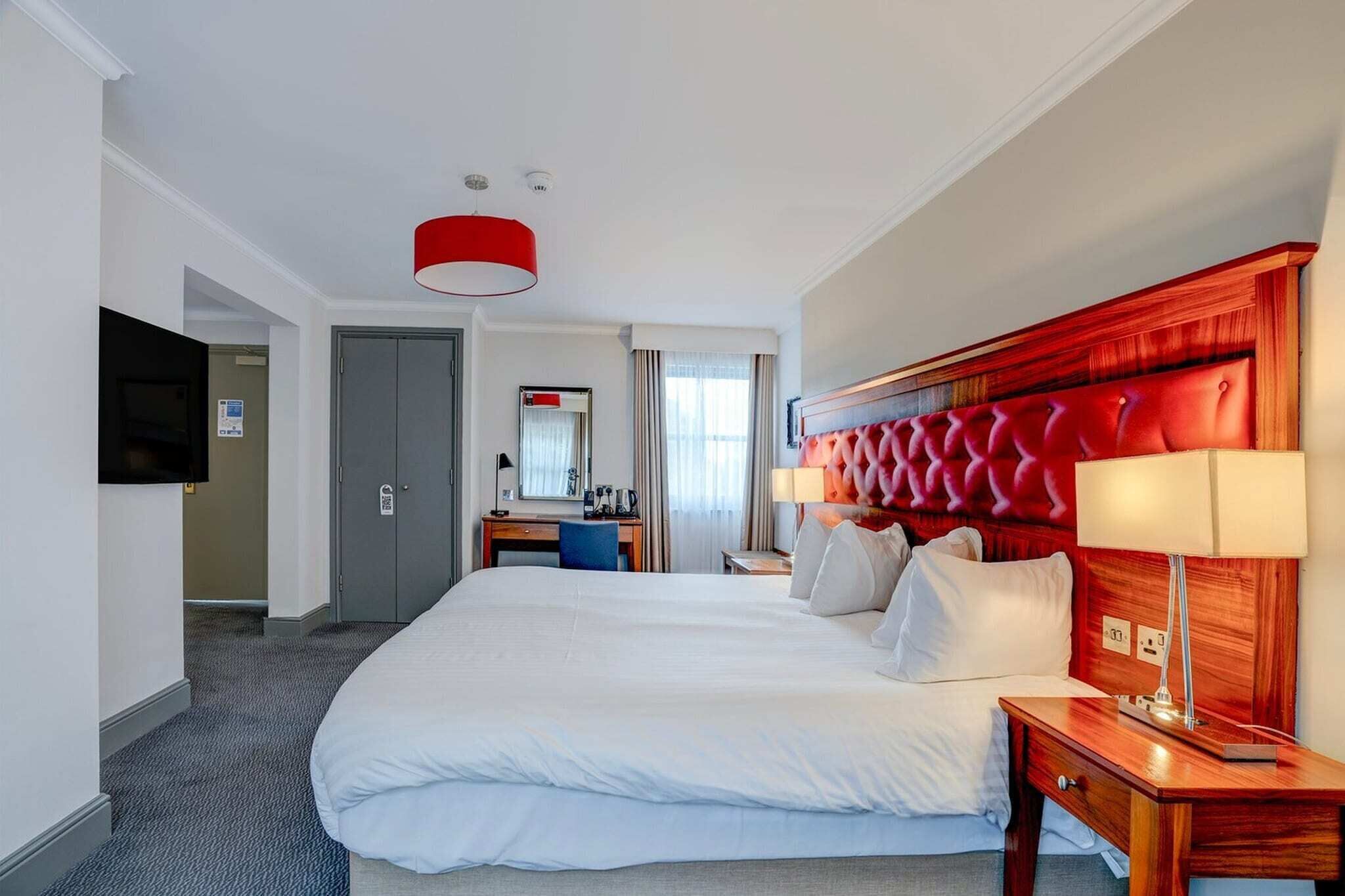 A hotel room with a large bed, a desk, a chair, a television, and a red accent wall.