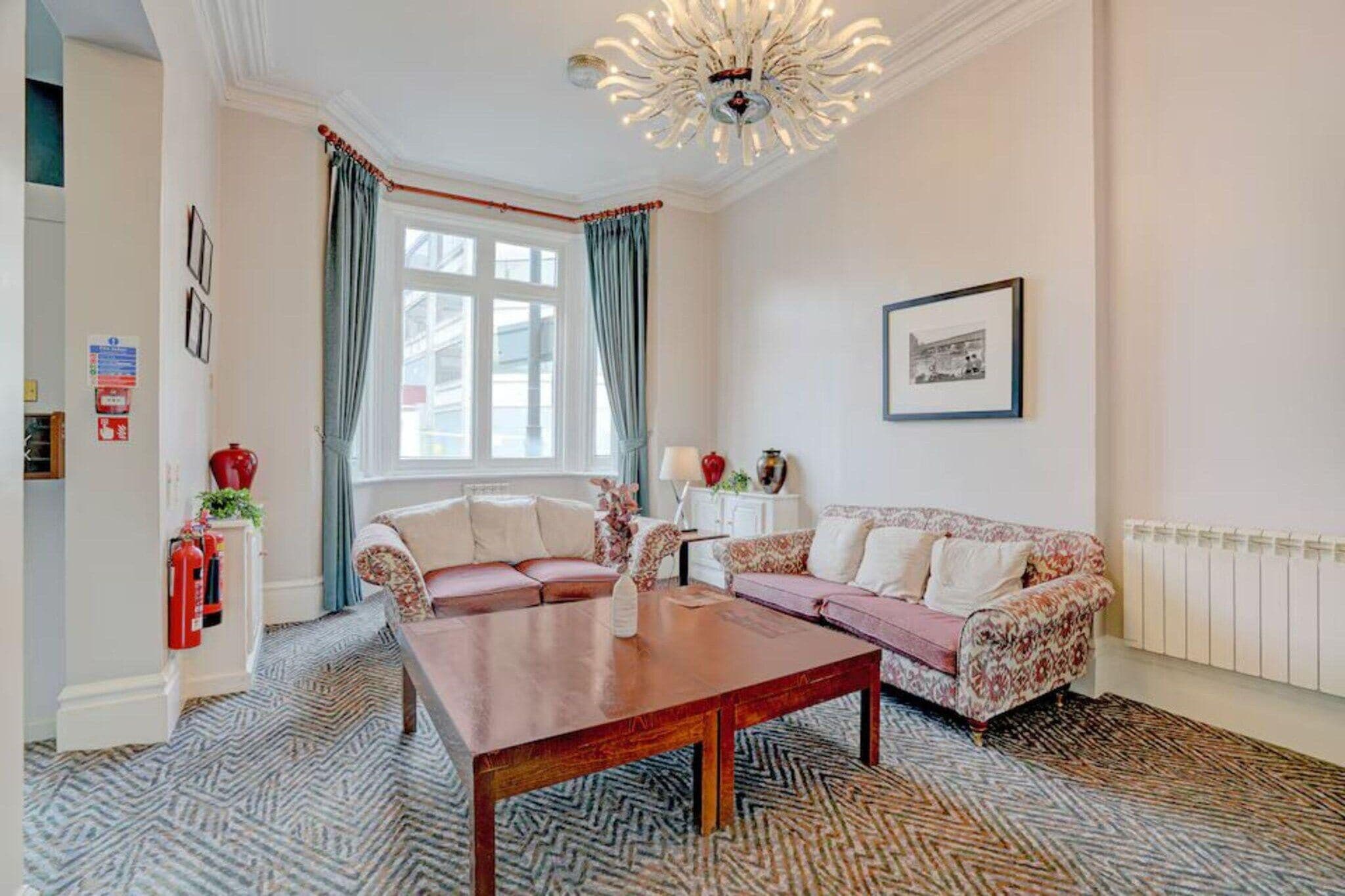 A room with a sofa set, a wooden coffee table, a patterned carpet, a window with curtains, and a wall-mounted picture.
