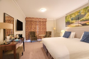 Habitación de hotel con cama, dos sillones, una mesita, un escritorio con lámpara, un espejo y un cuadro en la pared.