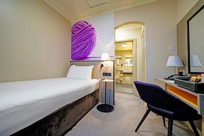 A hotel room with a bed, a desk, a chair, a lamp, a wall-mounted mirror, and a bathroom visible through an archway. - Standard Single Room (Exeter)