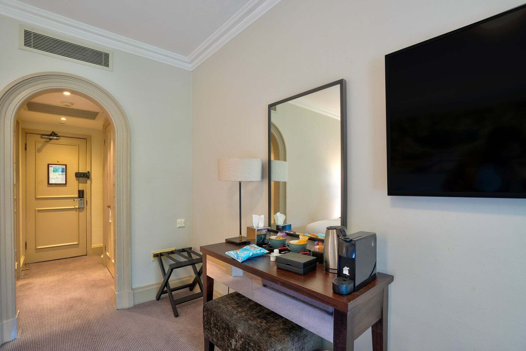 Ett hotellrum med ett skrivbord, en stor spegel, en platt-TV, en lampa och en kaffebryggare.