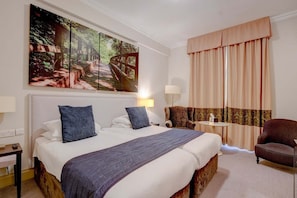 Habitación de hotel con una cama grande, dos almohadas, una mesita de noche, una lámpara, una silla, una pequeña mesa y un cuadro grande en la pared.