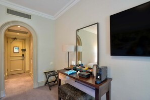 A hotel room with a desk, a large mirror, a flat-screen TV, a lamp, and a coffee maker.