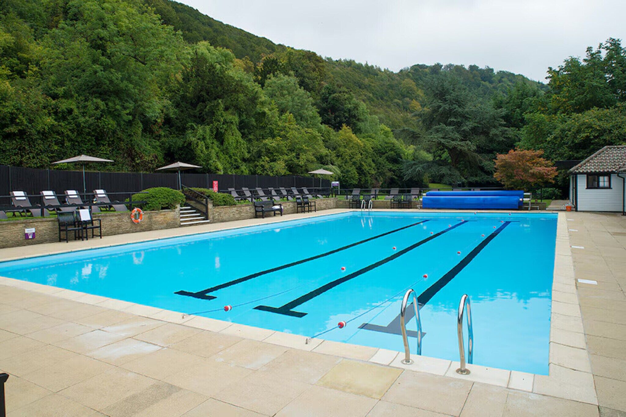 A swimming pool with multiple lanes, surrounded by lounge chairs and umbrellas, set against a backdrop of trees and hills.
