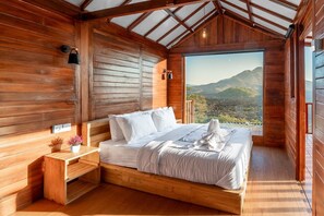 A wooden cabin bedroom with a bed, bedside table, and a view of a mountainous landscape. - Kamani Cabin (Bali)