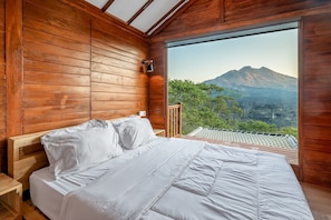 A wooden cabin bedroom with a bed, a nightstand, and a view of a mountain. - Kamani Cabin (Bali)
