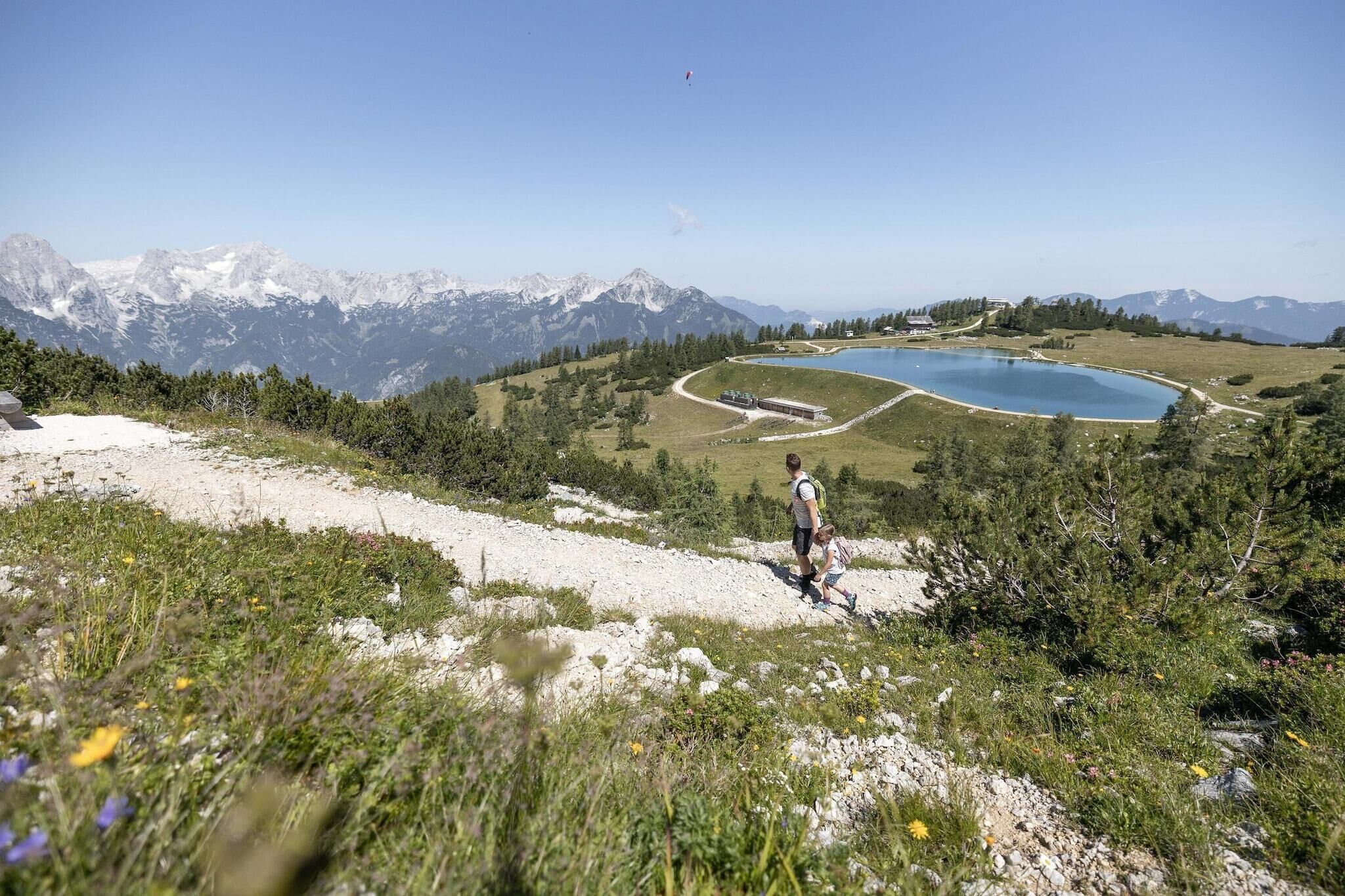 Un paisaje montañoso con un lago, una persona caminando y un cielo despejado.