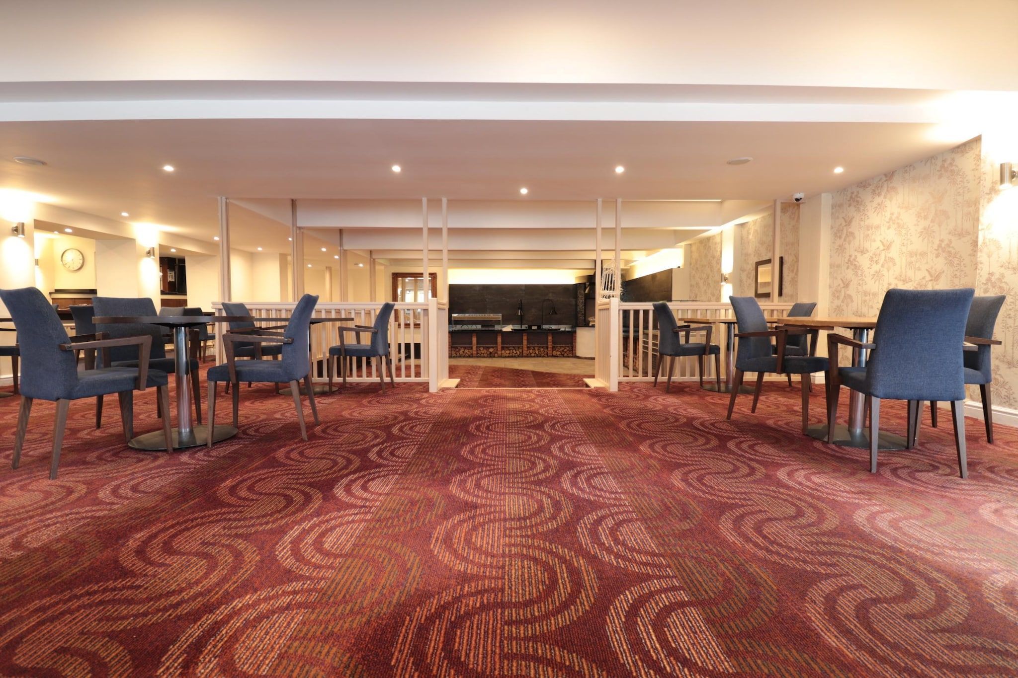 Un hall d’hôtel avec un tapis rouge à motifs, plusieurs tables et chaises, un comptoir de réception et une horloge au mur.