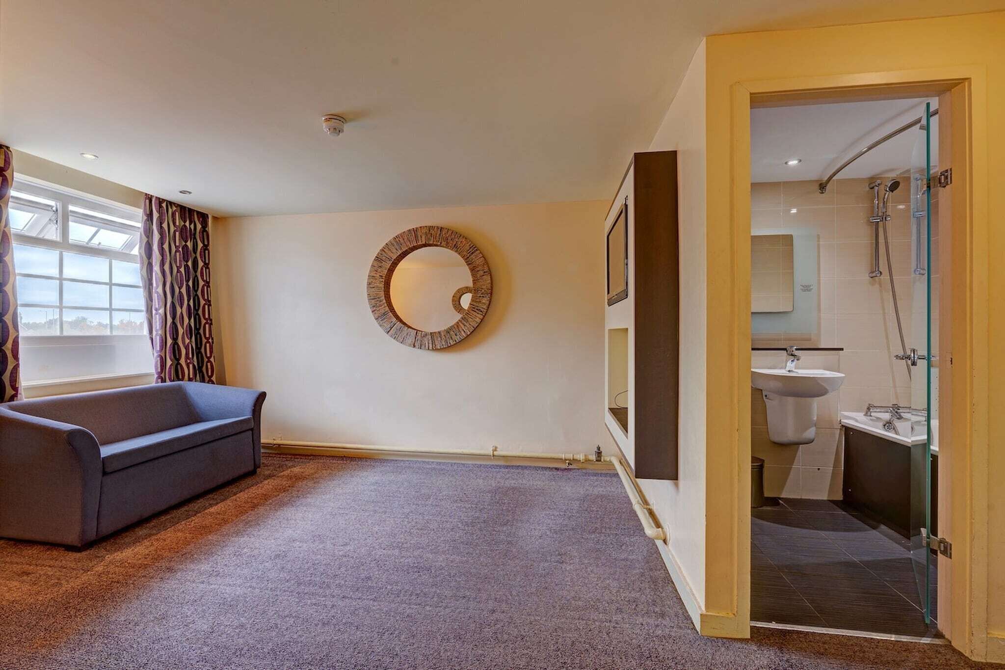 Een kamer met een bank, een ronde wanddecoratie, een glazen deur naar een badkamer en een raam met gordijnen.