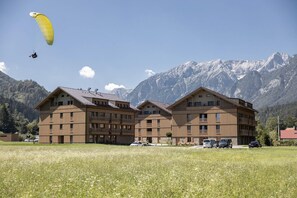 A multi-story building with balconies, a field in the foreground, and mountains in the background.