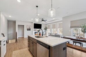 Une cuisine moderne dotée d’un îlot central, d’appareils électroménagers en acier inoxydable, de luminaires suspendus et offrant une vue sur l’extérieur.