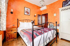 A bedroom with an orange wall, a bed with a patterned blanket, a wooden nightstand, a white wardrobe, and a chandelier.