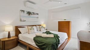 Et pænt opredt soveværelse med seng, sengeborde med lamper, en fastgjort aircondition, ventilator i loftet, et maleri og en trædør.
