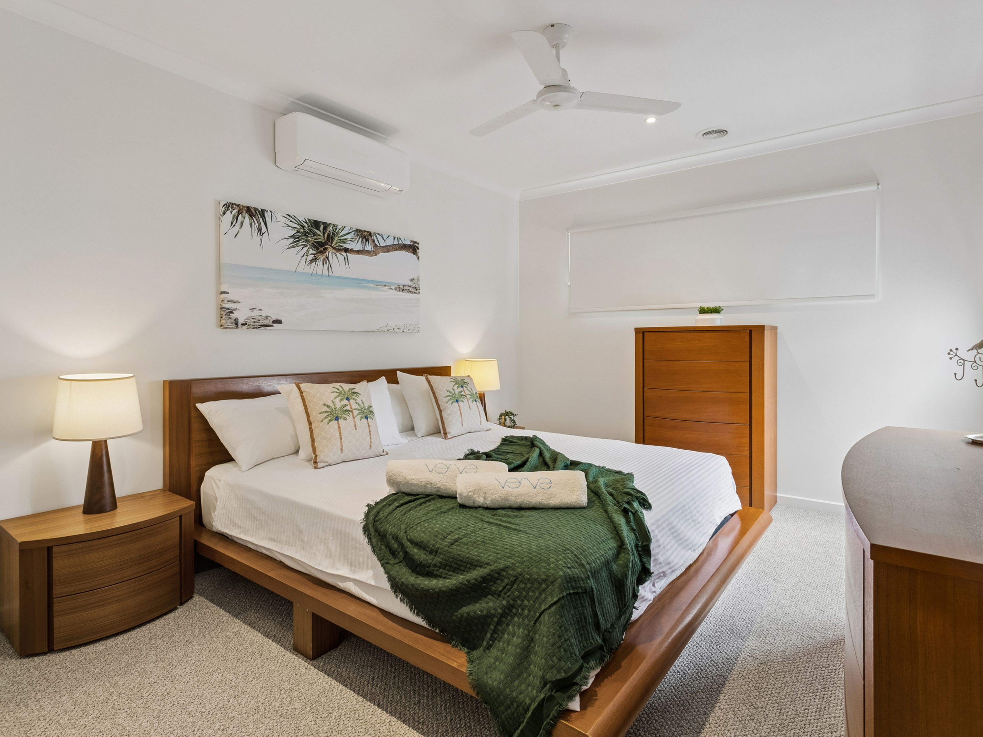 Una habitación de hotel con cama, mesitas de noche, ventilador de techo, equipo de aire acondicionado, un cuadro con una playa y un armario de madera.