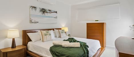 Una habitación de hotel con cama, mesitas de noche, ventilador de techo, equipo de aire acondicionado, un cuadro con una playa y un armario de madera.