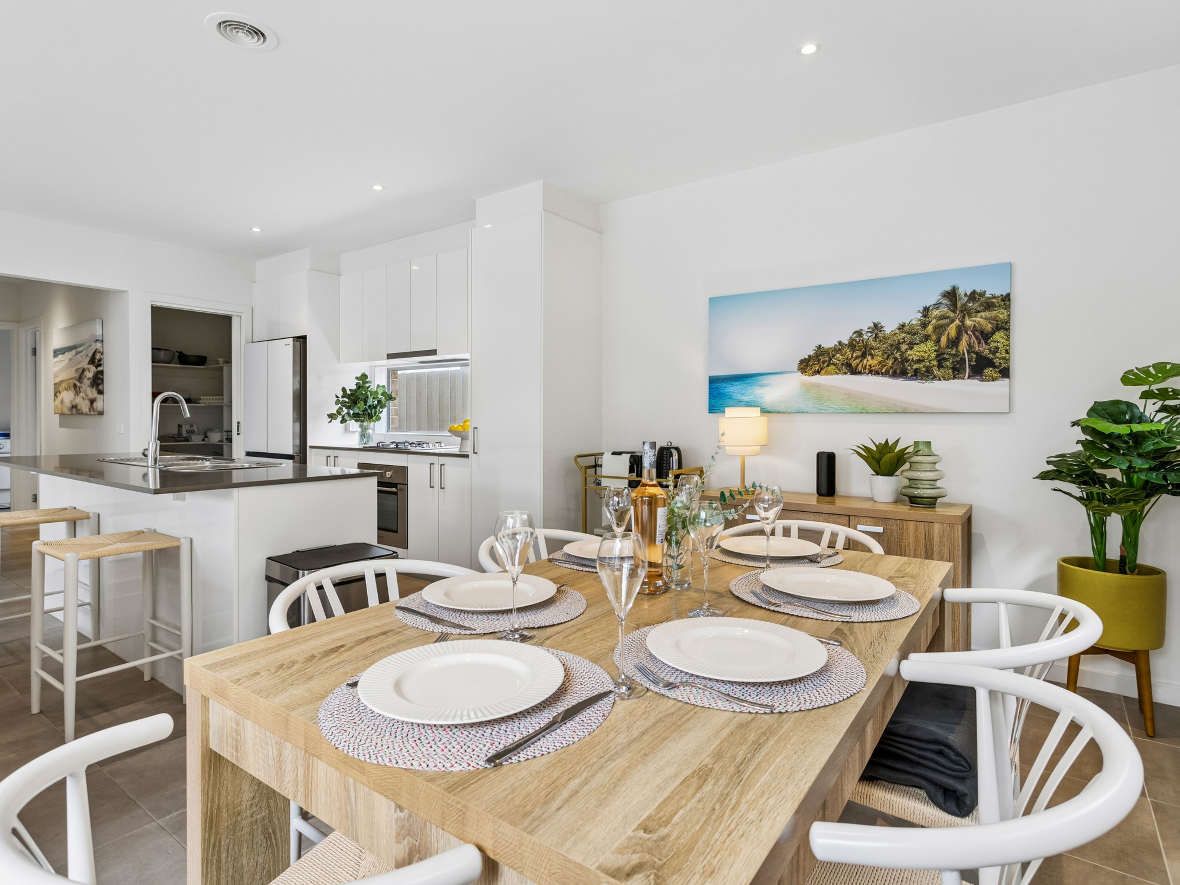 A modern kitchen with white cabinets, a dining area with a wooden table set for four, and a painting of a beach scene on the wall.