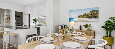 A modern kitchen with white cabinets, a dining area with a wooden table set for four, and a painting of a beach scene on the wall.