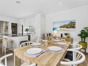 Una cucina moderna con mobili bianchi, un'area pranzo con tavolo in legno apparecchiata per quattro persone e un quadro raffigurante una spiaggia sulla parete.