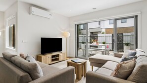 En moderne stue med et fladskærms-tv, møbler i træ og en stor skydedør, der fører ud til en udendørs terrasse.