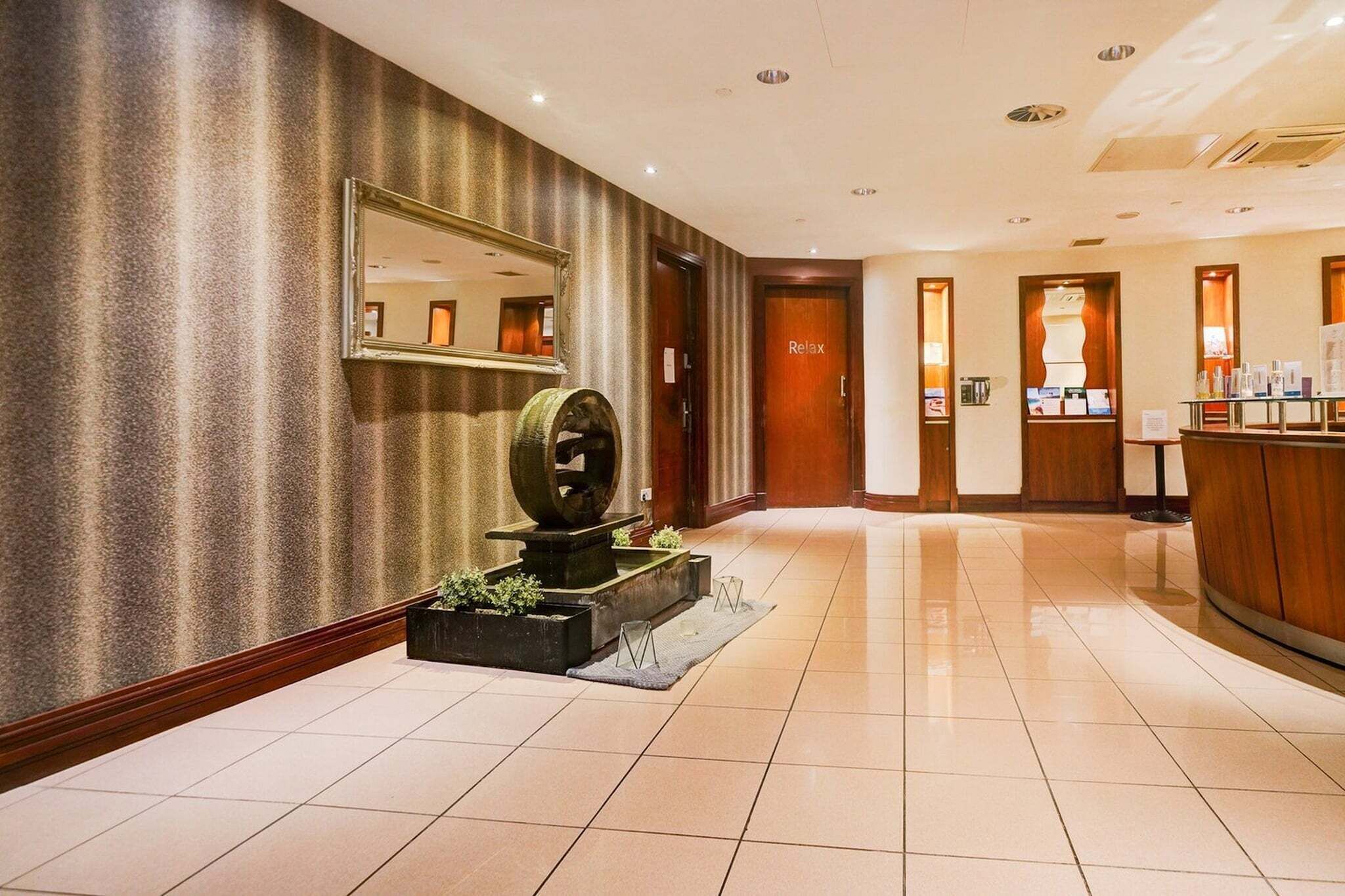 A hotel lobby with a reception desk, a 'Relax' sign, a large mirror, and a decorative fountain.