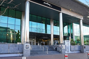 Une entrée d’hôtel moderne, avec de grandes fenêtres en verre, un escalier menant au hall d’accueil et une enseigne bien visible indiquant « Sunday Hotel ».