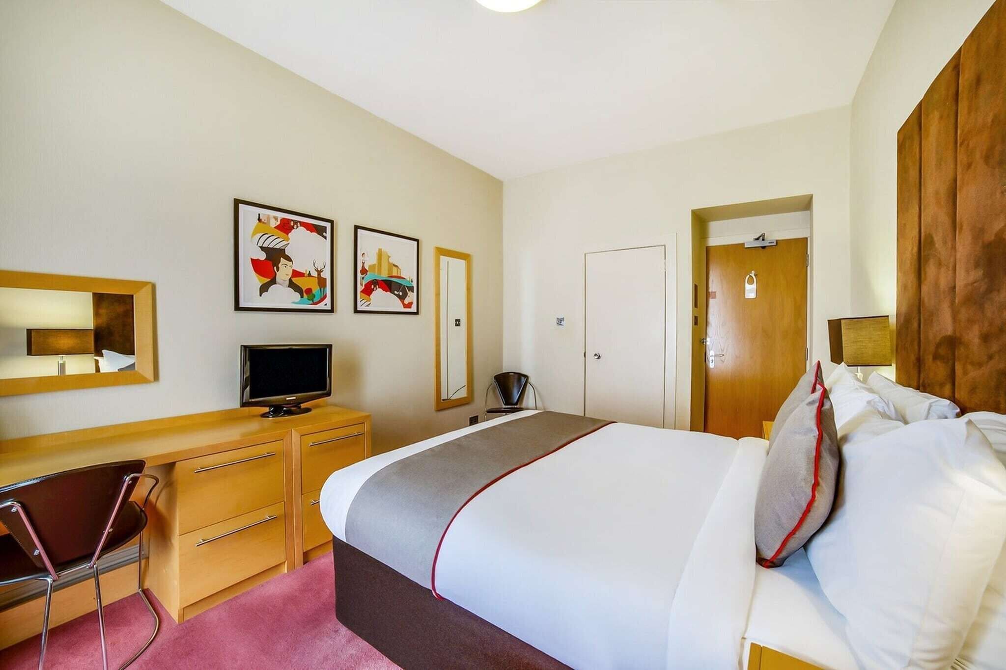 A hotel room with a bed, a desk with a computer, a chair, a television, and artwork on the walls.