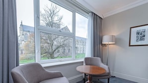 Une pièce avec une grande fenêtre, une chaise, une petite table ronde, une lampe et un tableau encadré au mur.