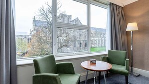 Une chambre d’hôtel avec une grande fenêtre, deux fauteuils verts, une petite table ronde et un lampadaire.