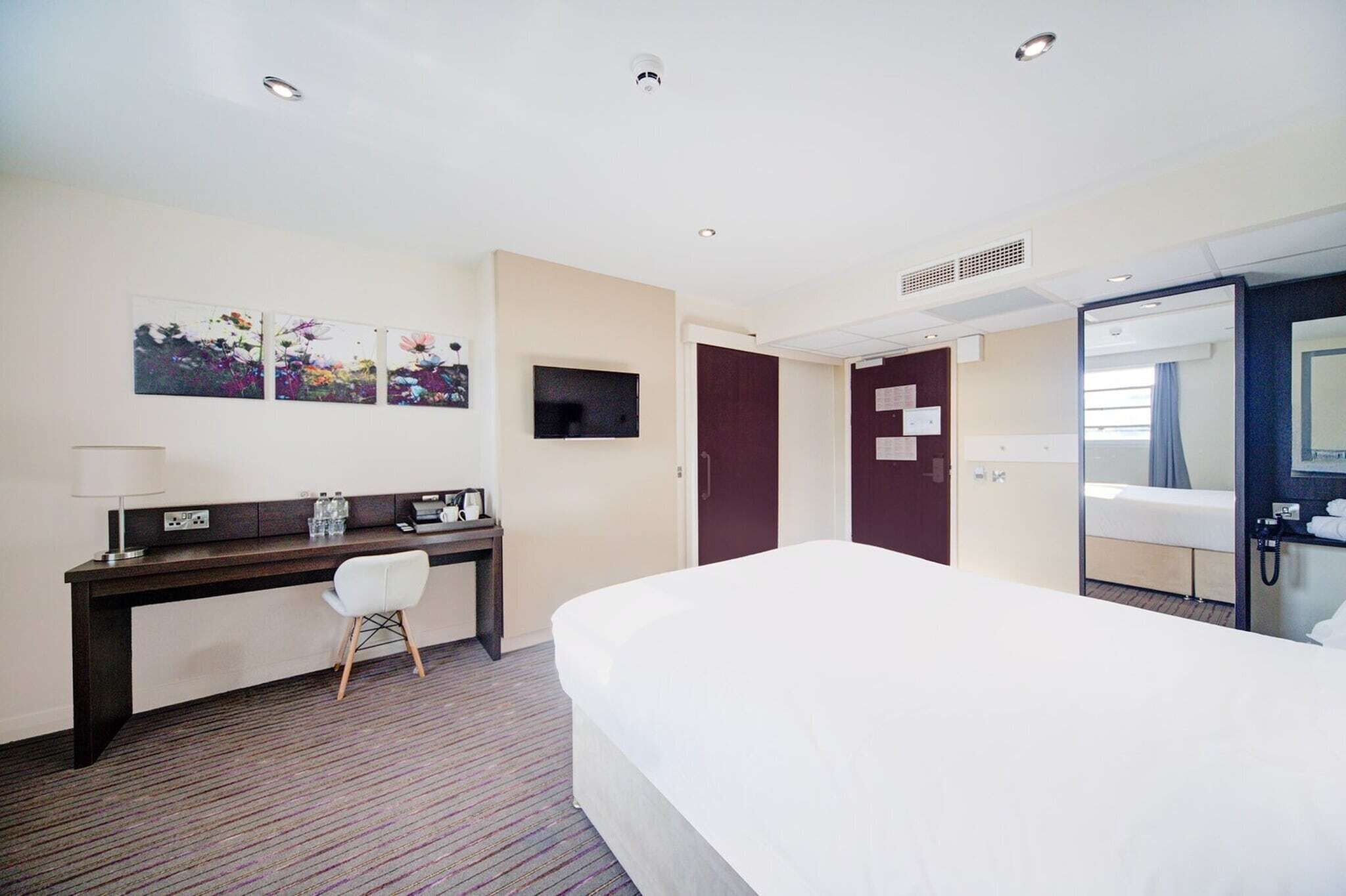 A hotel room with a bed, desk, chair, TV, and a bathroom mirror.