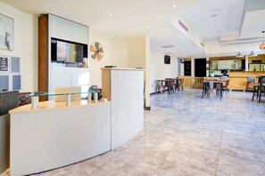 En moderne hotellobby med reception, siddeområde og et køkken i baggrunden.