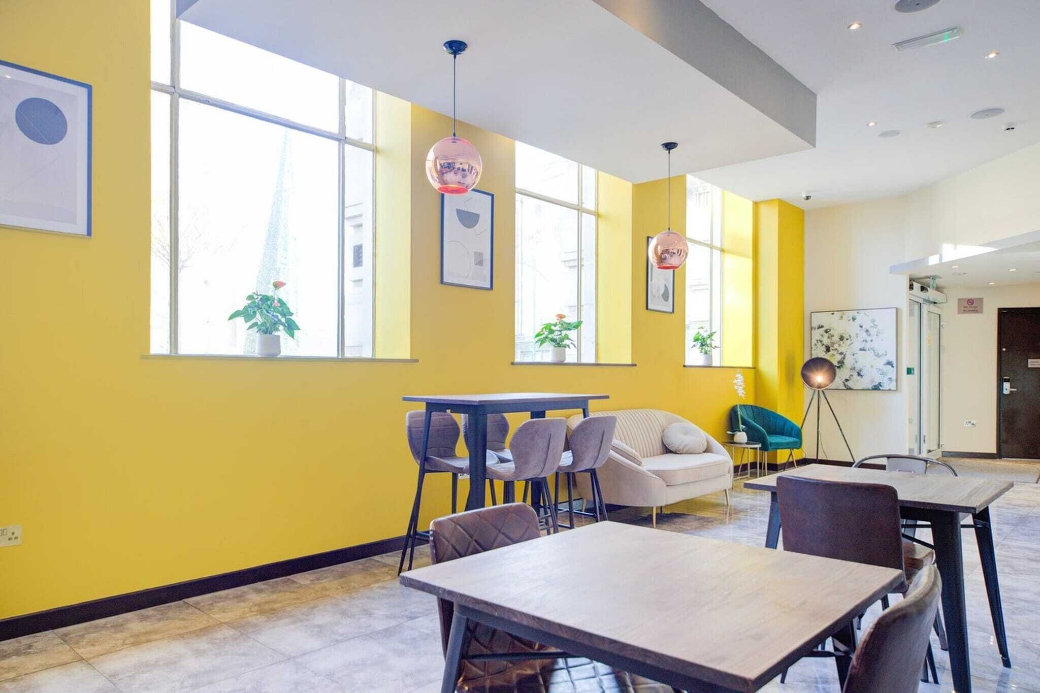 A modern interior with yellow walls, large windows, a dining area with tables and chairs, and a sofa.