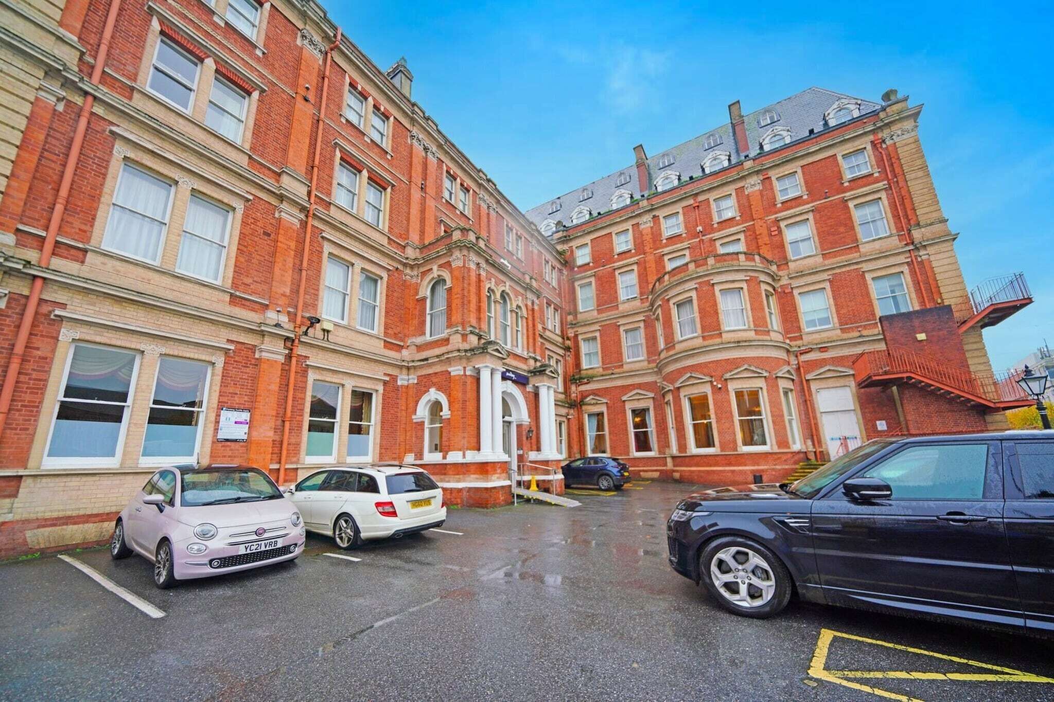 A multi-story building with a red brick facade, a parking lot with several cars, and a clear blue sky.