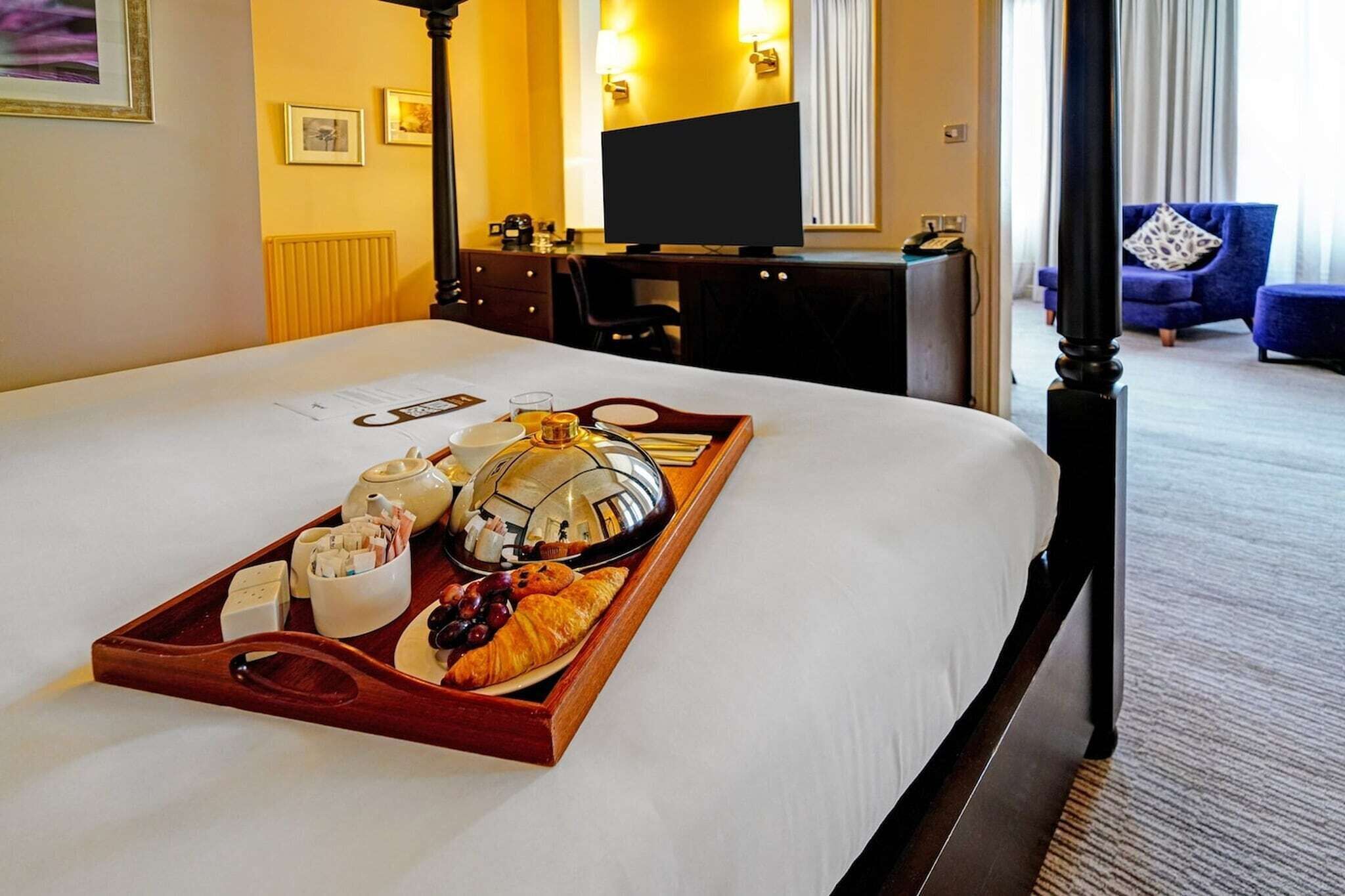 A hotel room with a neatly made bed, a tray with breakfast items, a desk with a computer, and a TV.