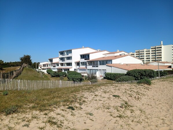 A coastal residential area with modern white buildings, a sandy beach, and a wooden fence.