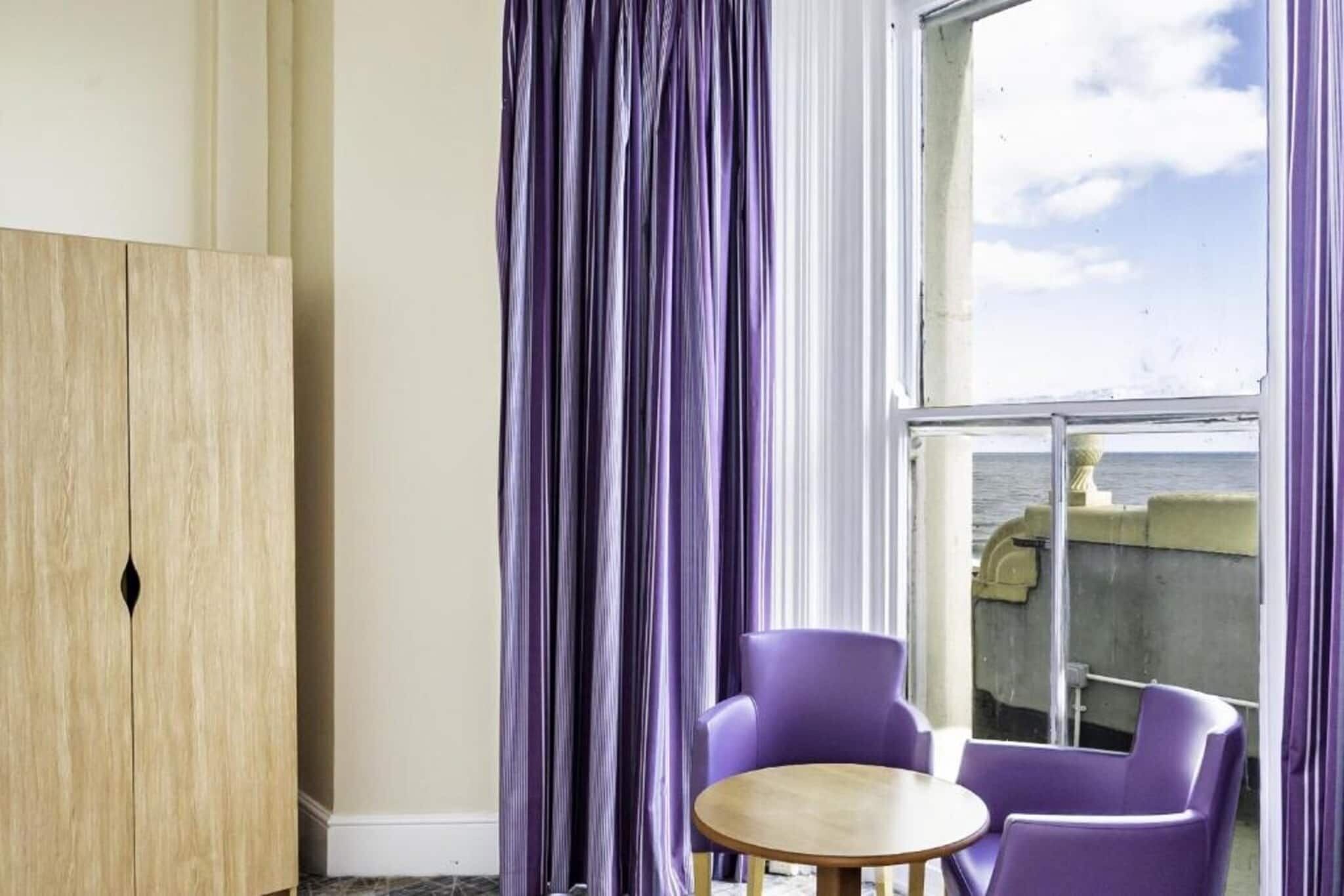 A room with a wooden wardrobe, purple chairs, a small round table, and a large window with a view of the sea.