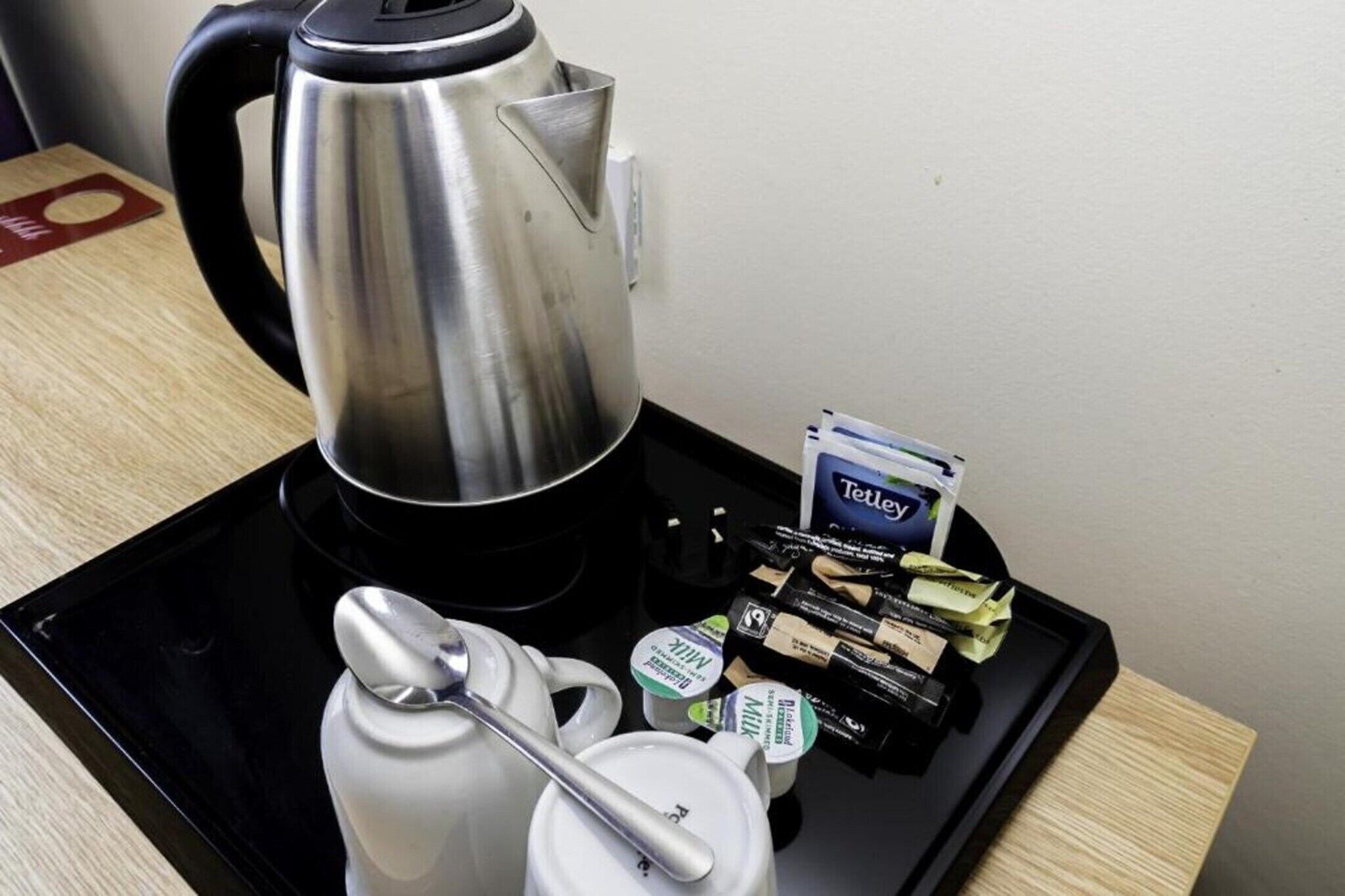 Ruostumattomasta teräksestä valmistettu vedenkeitin, valkoinen teekannu, lusikka sekä tarjotinalusta, jossa on Tetley-teepusseja ja maitotölkkejä.
