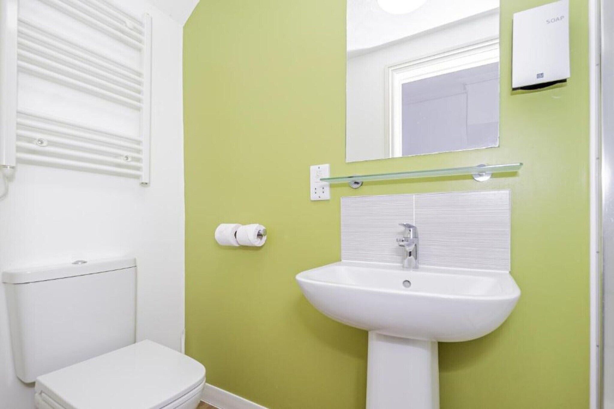 Een badkamer met een wit toilet, een witte wastafel, een glazen plank, een spiegel en een zeepdispenser.