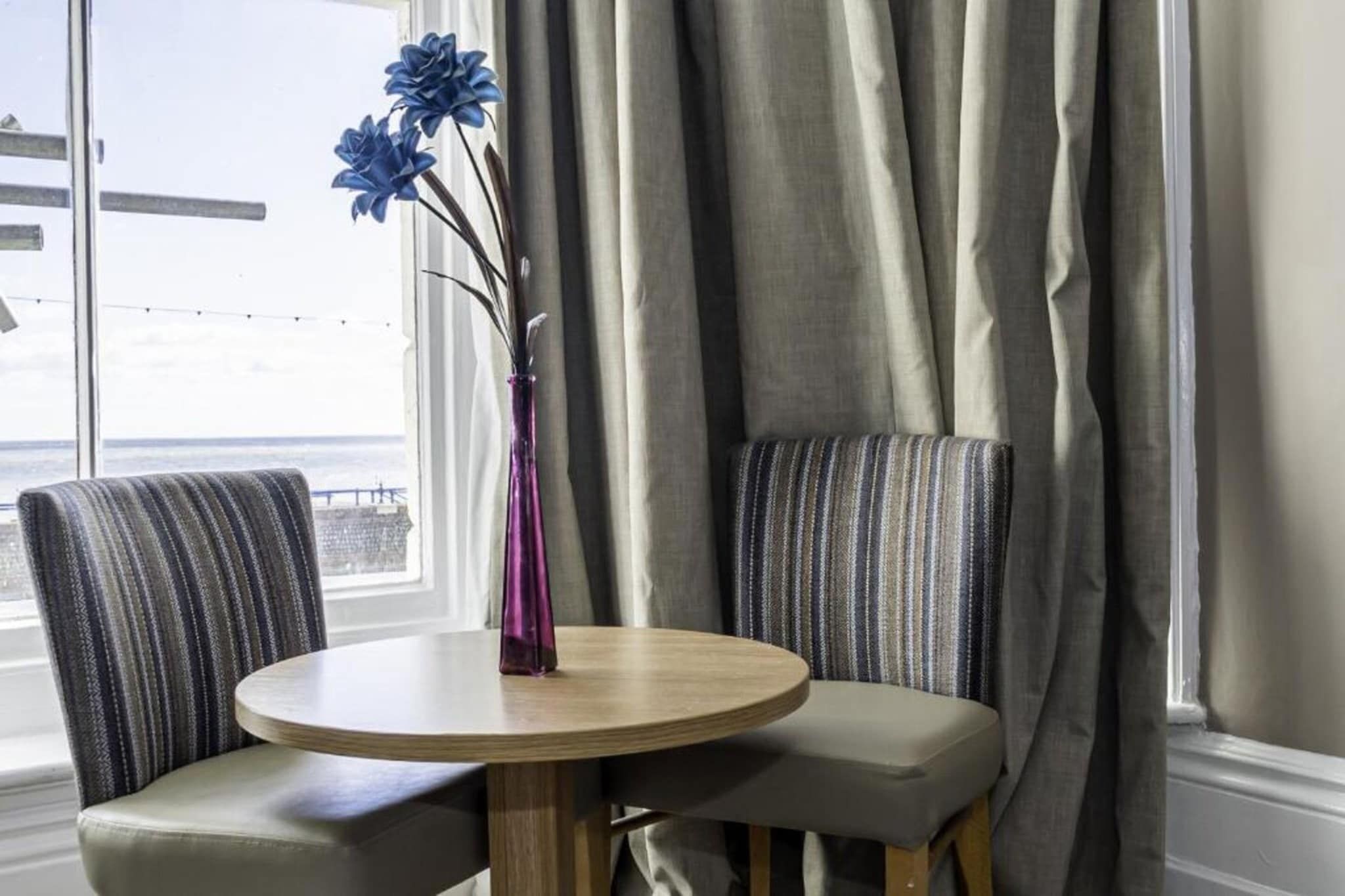 Une table ronde, deux chaises rembourrées, un vase de fleurs et une vue sur la mer par une fenêtre ornée de rideaux.